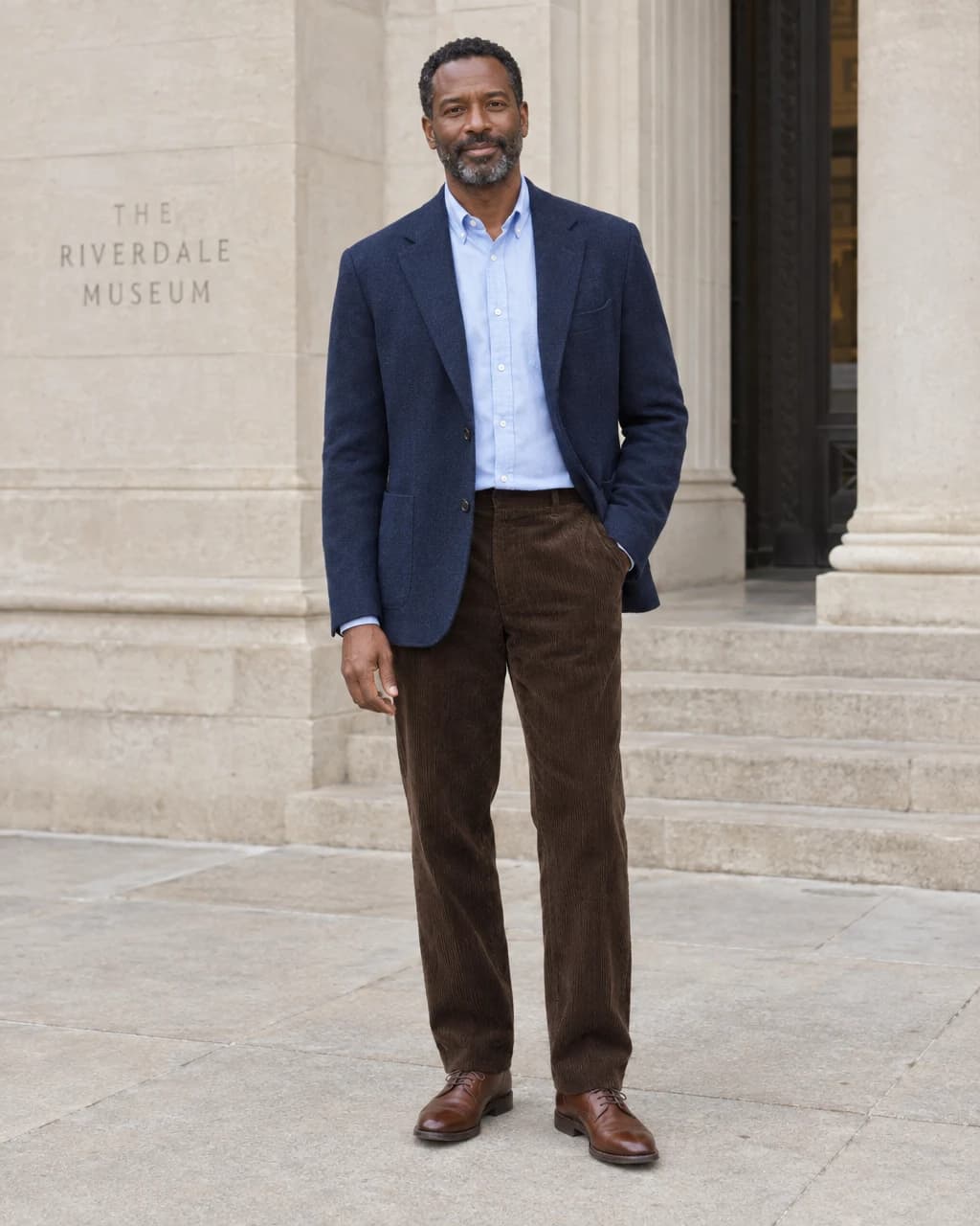 Man wearing a navy sport coat, pale blue shirt, dark brown corduroy trousers and brown leather lace-up shoes outside a limestone entrance