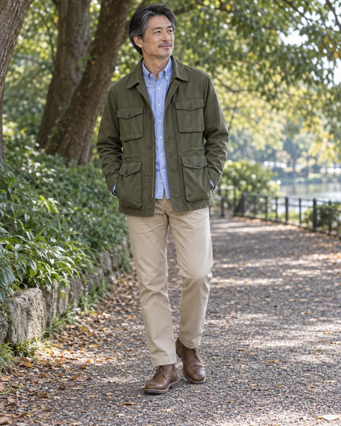 Middle-aged East Asian man wearing an olive four-pocket field jacket over a pale blue Oxford with stone chinos and brown chukka boots in a park
