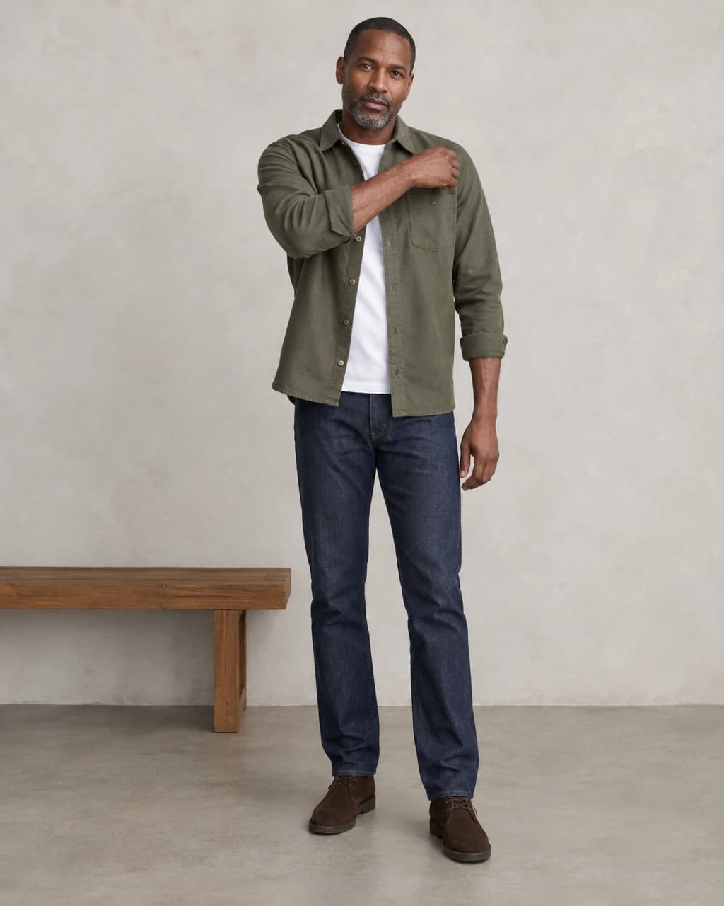 Man checking shoulder movement in an olive overshirt over a white tee, dark jeans and suede boots beside a wooden bench