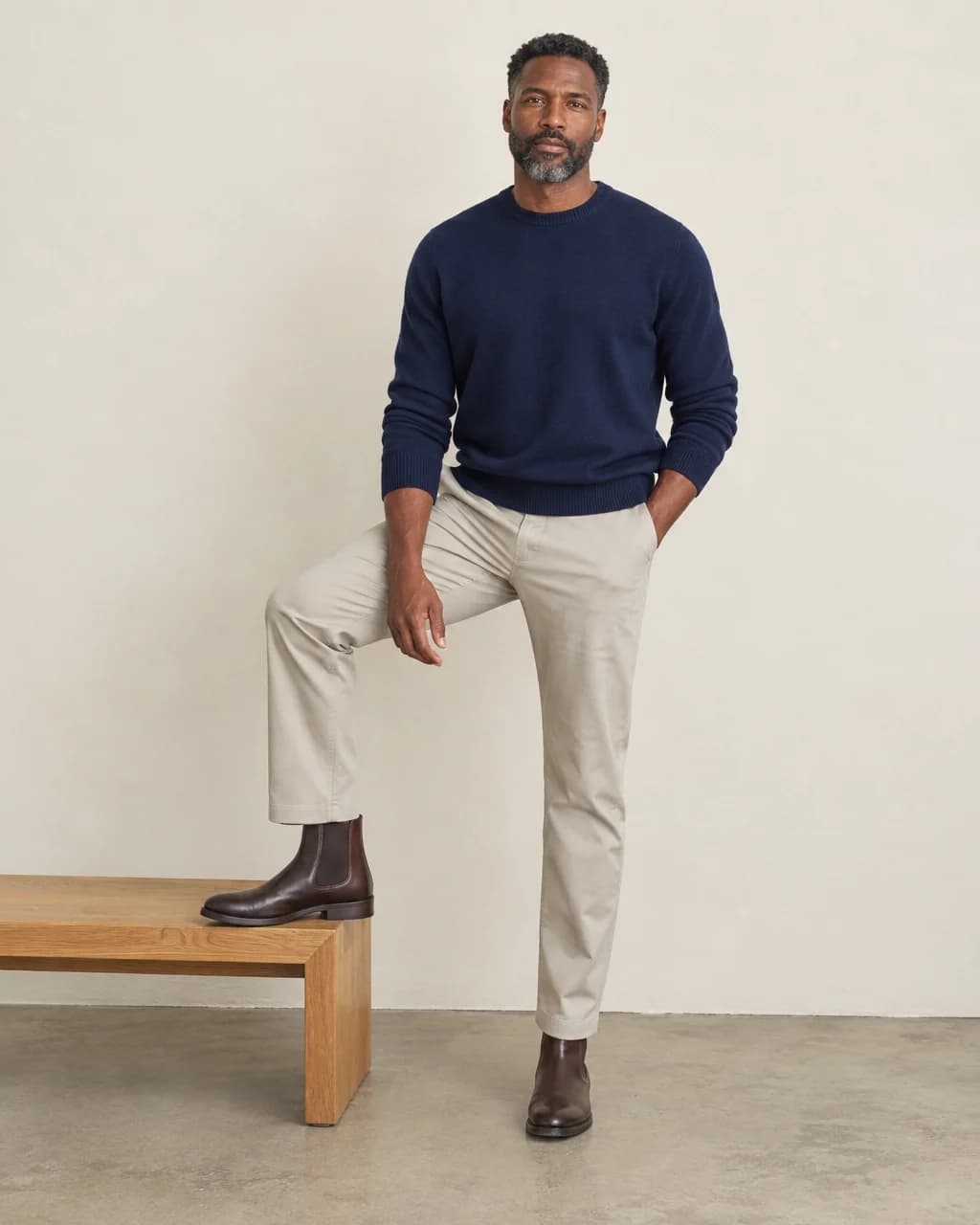 Man in navy sweater and stone chinos comparing a trouser hem against brown Chelsea boots beside a wooden bench