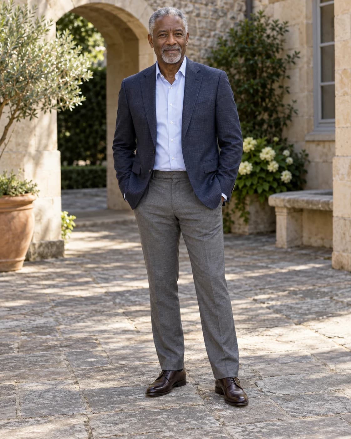 Mature Black man in a navy blazer, pale blue shirt, gray trousers and dark brown shoes in a sunlit courtyard