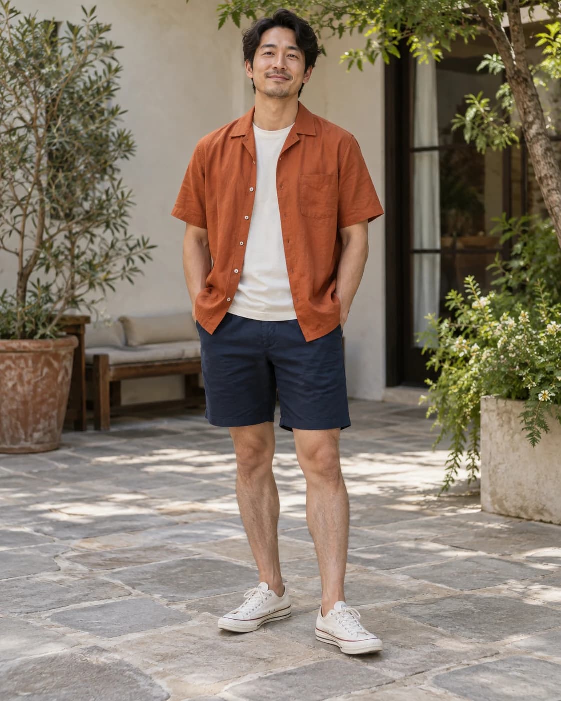 Adult East Asian man wearing a burnt-orange camp shirt over a cream tee with navy shorts and off-white trainers