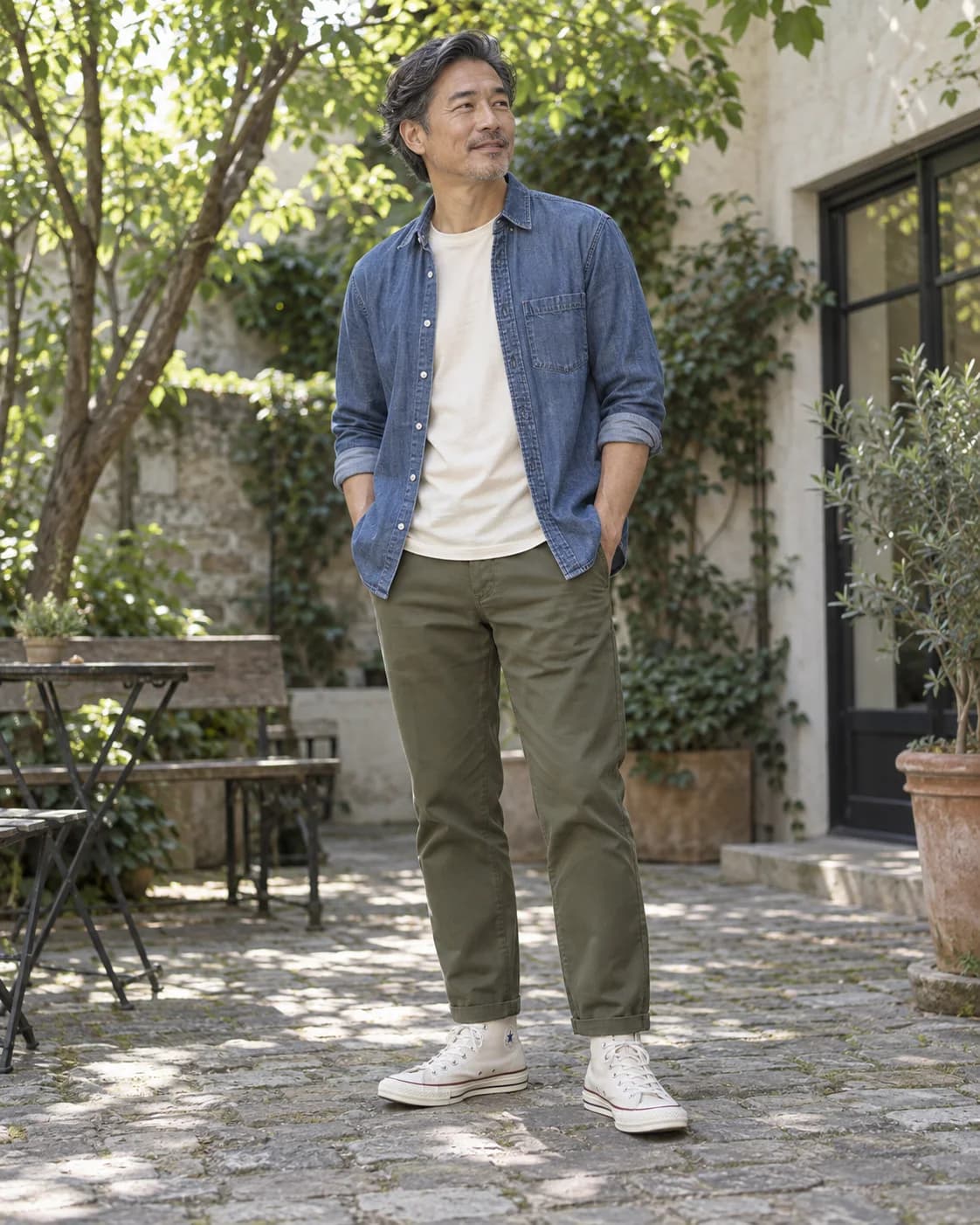 Adult East Asian man in natural ivory high-top canvas sneakers, olive chinos, blue denim shirt and cream tee in a leafy courtyard