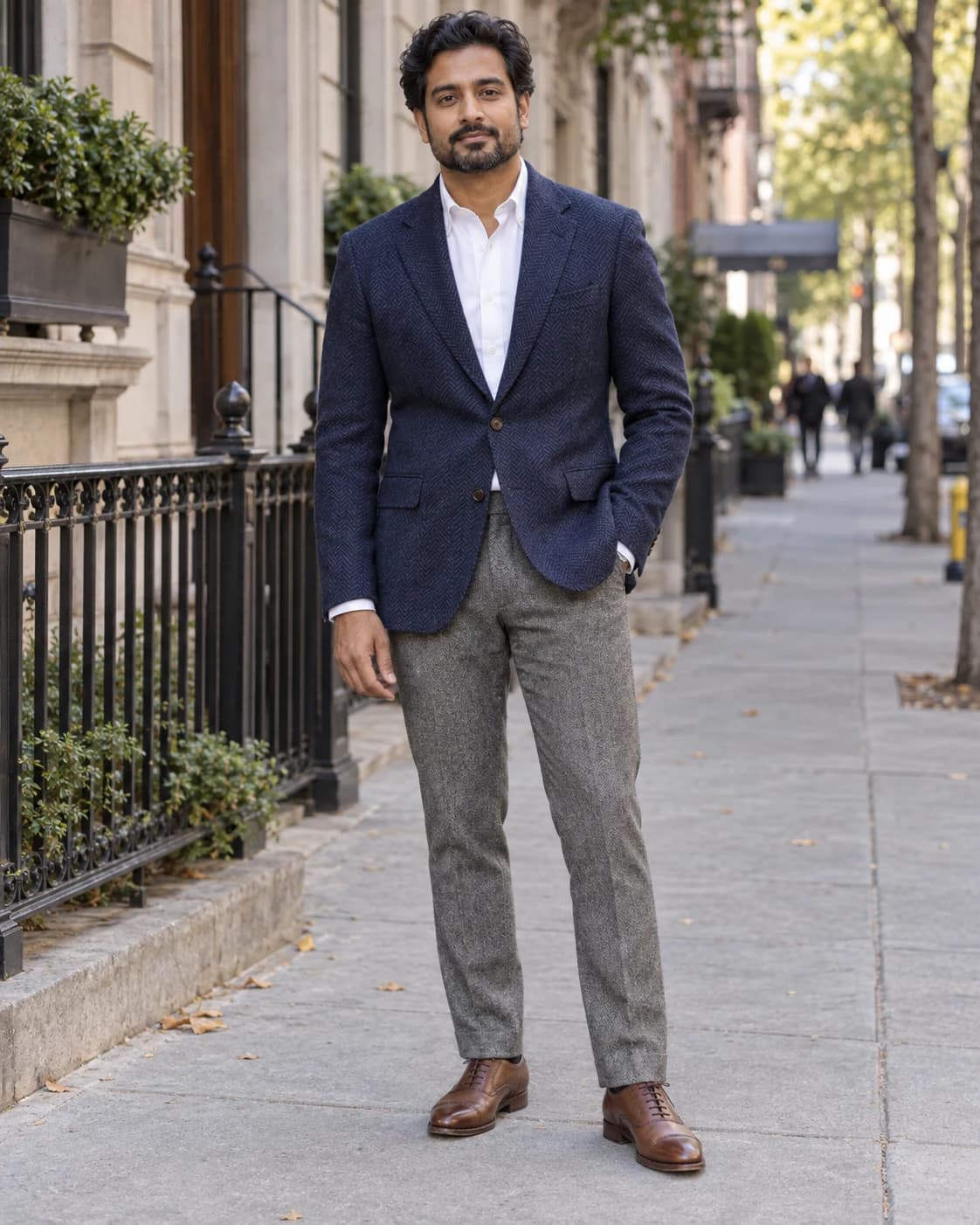 Adult South Asian man in a navy herringbone blazer, white Oxford, gray trousers and brown cap-toe shoes
