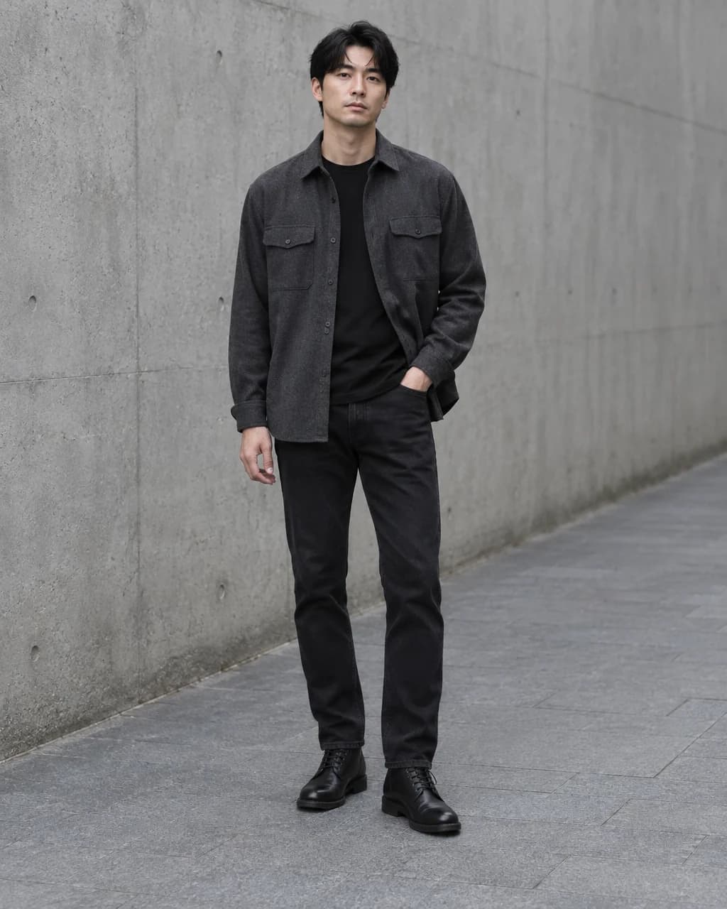 Man in a charcoal overshirt, black T-shirt, straight black jeans and black lace-up boots beside concrete