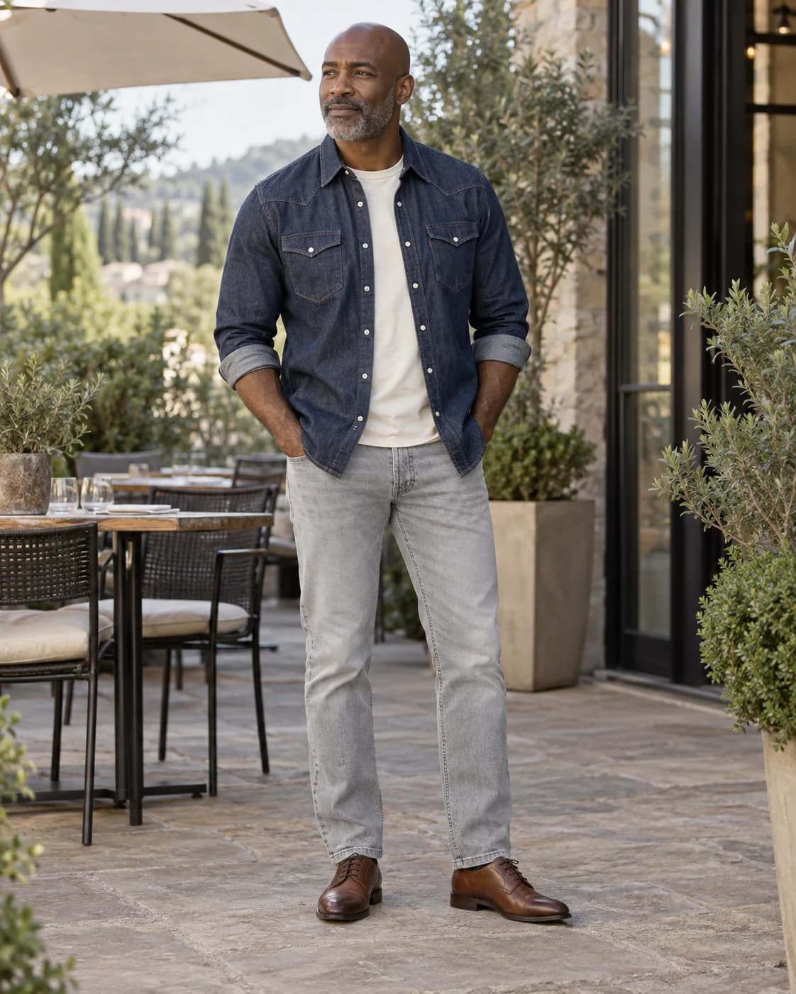 Adult Black man wearing an indigo Western denim snap shirt over a cream tee with light-gray jeans and brown cap-toe shoes at an outdoor lunch